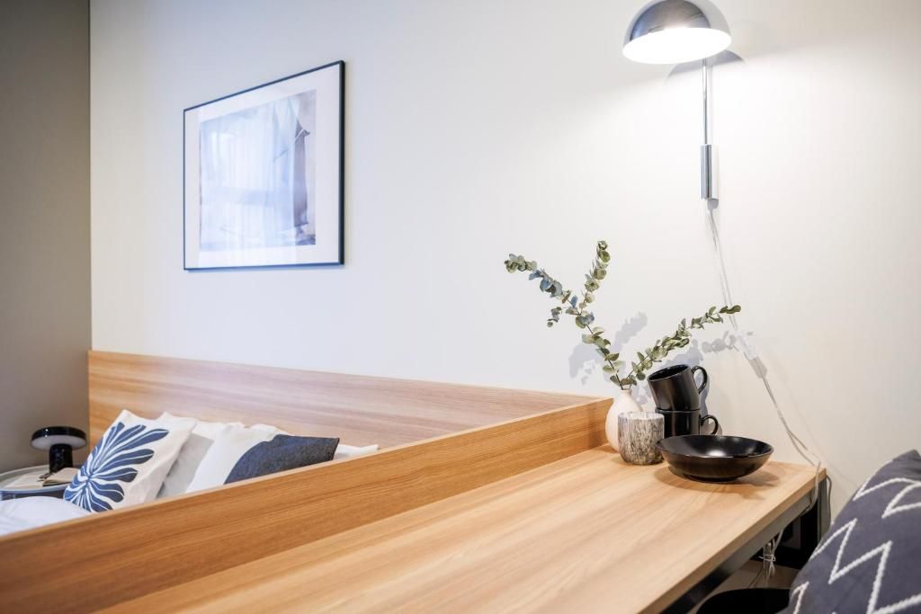 Quarto de hotel com cabeceira de madeira, mesa de cabeceira com abajur, quadro na parede e vaso com planta.
