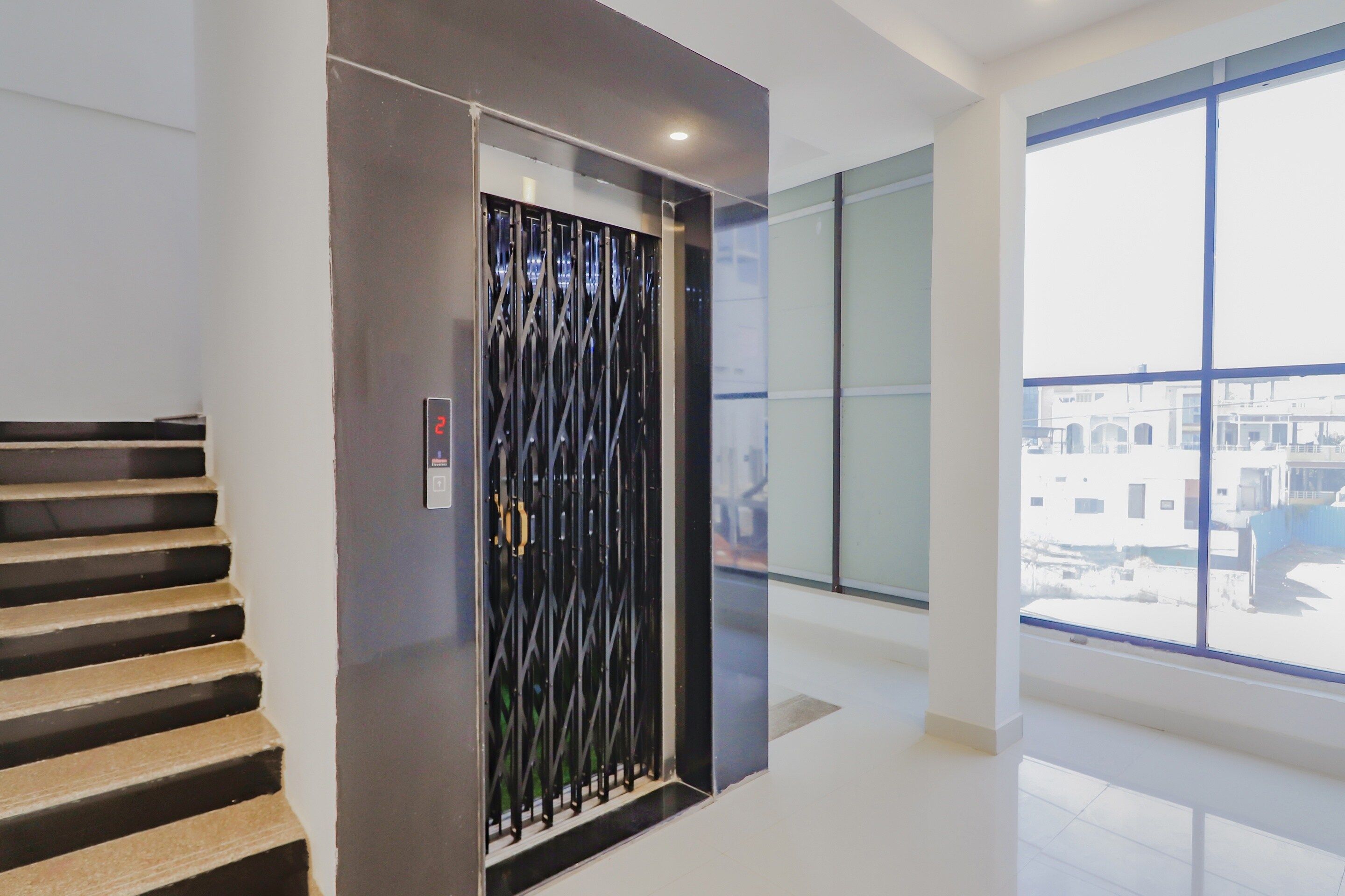 Un ascensor moderno con puerta metálica de diseño, una escalera con escalones de madera y un ventanal con vistas a edificios.