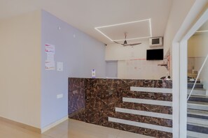Zona de recepción de un hotel con un mostrador de mármol, un ventilador de techo, un televisor de pantalla plana y una escalera que conduce a otro piso.