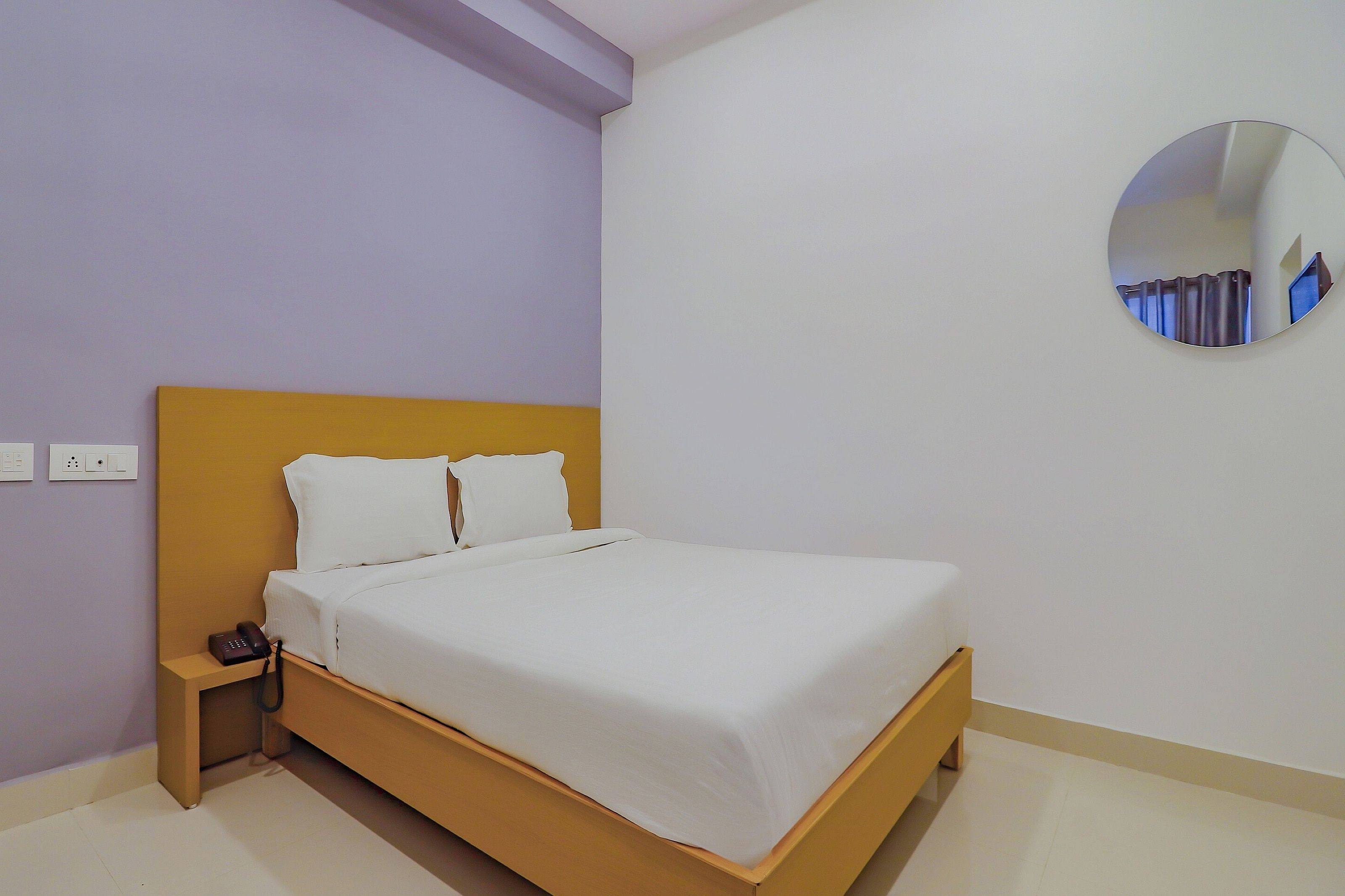 Una cama individual con estructura de madera y ropa de cama blanca en una habitación con paredes moradas claras y suelo blanco.