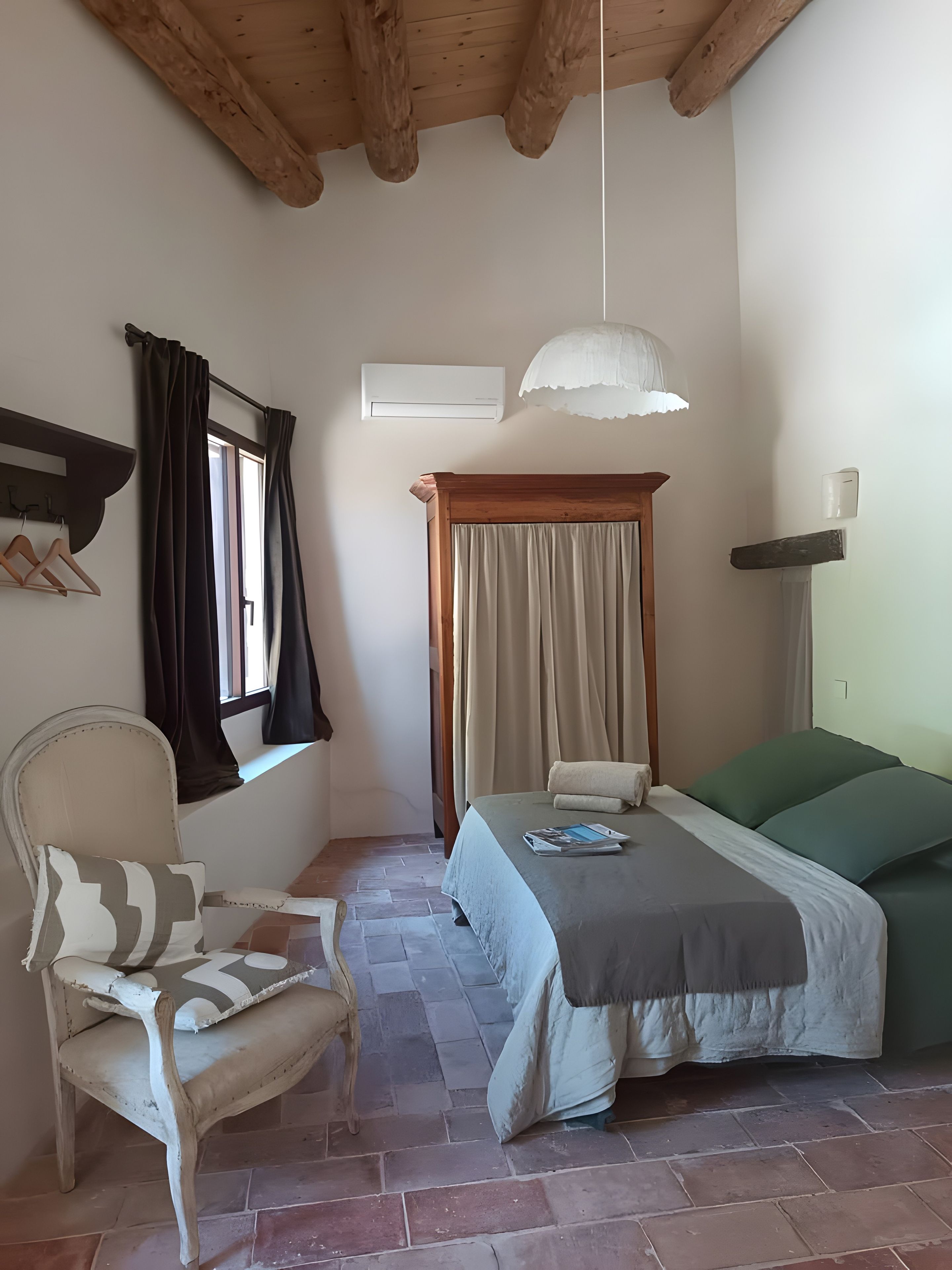 Ein Schlafzimmer mit einem Bett, einem Sessel, einem Fenster mit Vorhängen, einer Klimaanlage und einer Pendelleuchte.