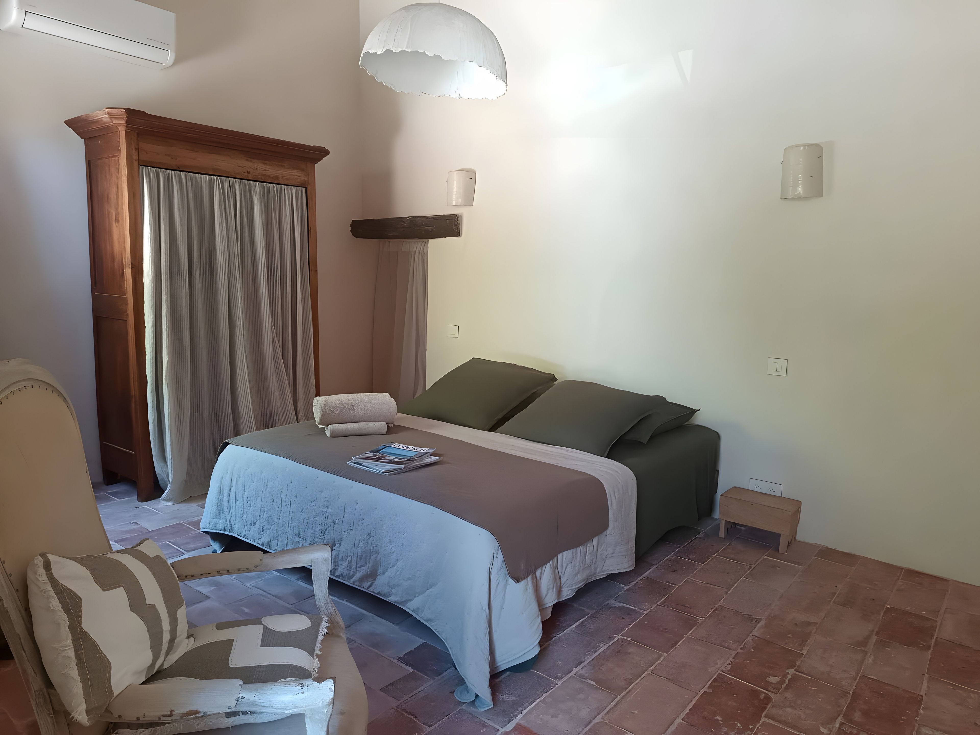 A bedroom with a bed, a chair, a bedside table, a window with curtains, and a wall-mounted air conditioner.