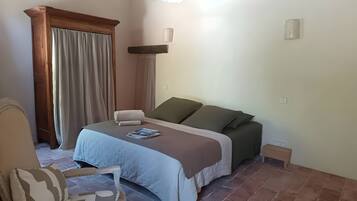 A bedroom with a bed, a chair, a bedside table, a window with curtains, and a wall-mounted air conditioner.