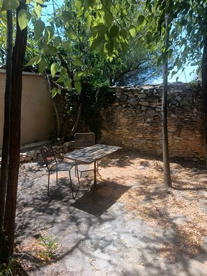 An outdoor patio with a table and chairs, surrounded by trees and a stone wall. - Studio 'Bëj-Gànnaar' with Air Conditioning and Garden - Luberon (Saint-Saturnin-lès-Apt)