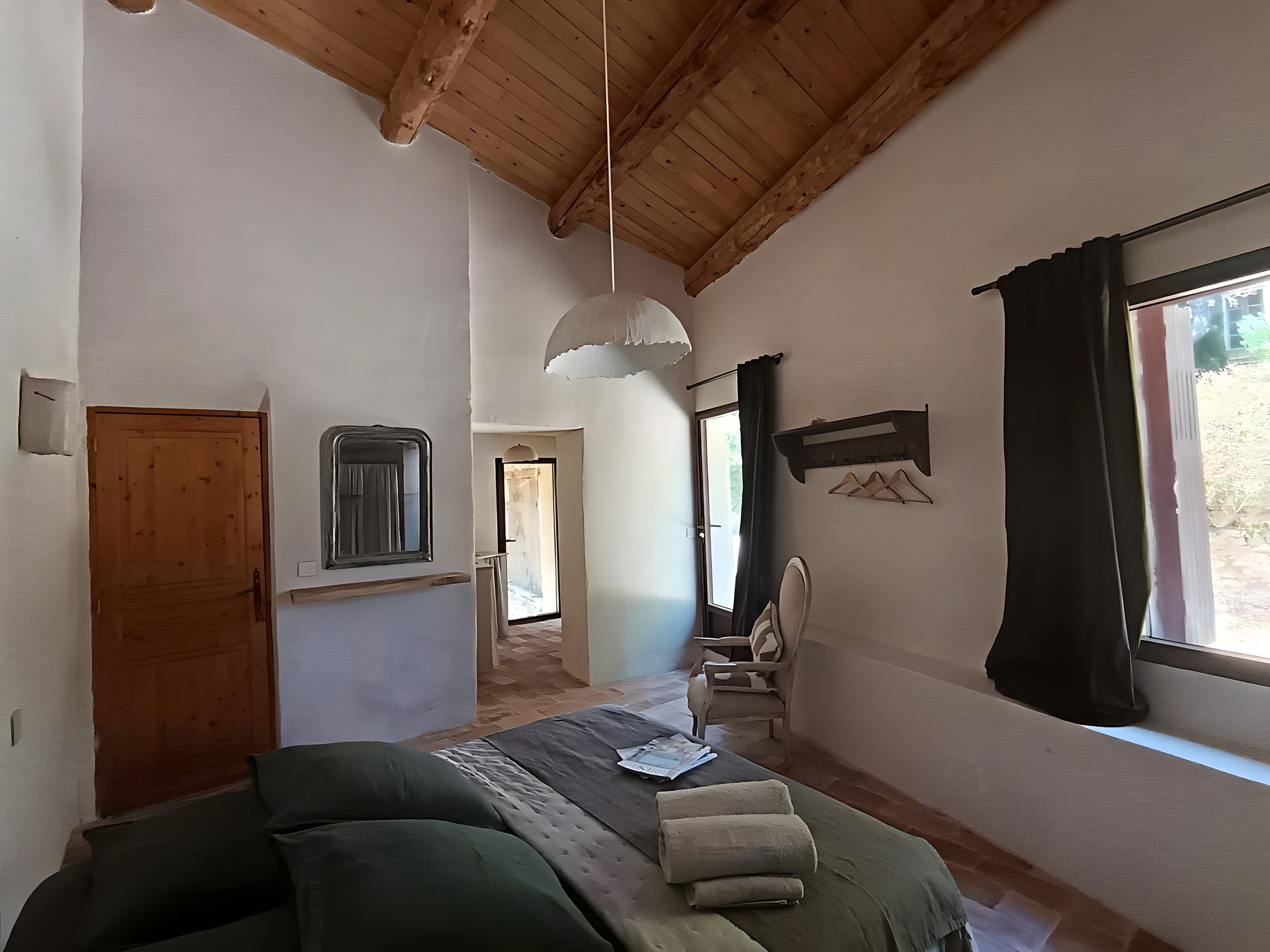 A room with a bed, a chair, a window with curtains, a hanging light fixture, and a wooden door.