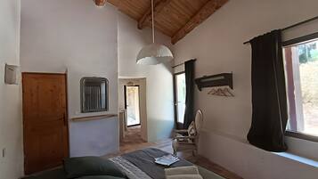 A room with a bed, a chair, a window with curtains, a hanging light fixture, and a wooden door.