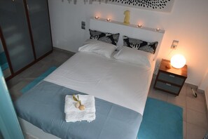 A neatly made bed with a white and blue bedspread, a white towel with seashells, and a bedside table with a lamp. - Clio's Beach House - Deluxe Beach front property (Ormos Korthiou)
