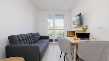Un salon moderne comprenant un canapé, une table à manger avec des chaises, un téléviseur fixé au mur et une fenêtre donnant sur l’extérieur.