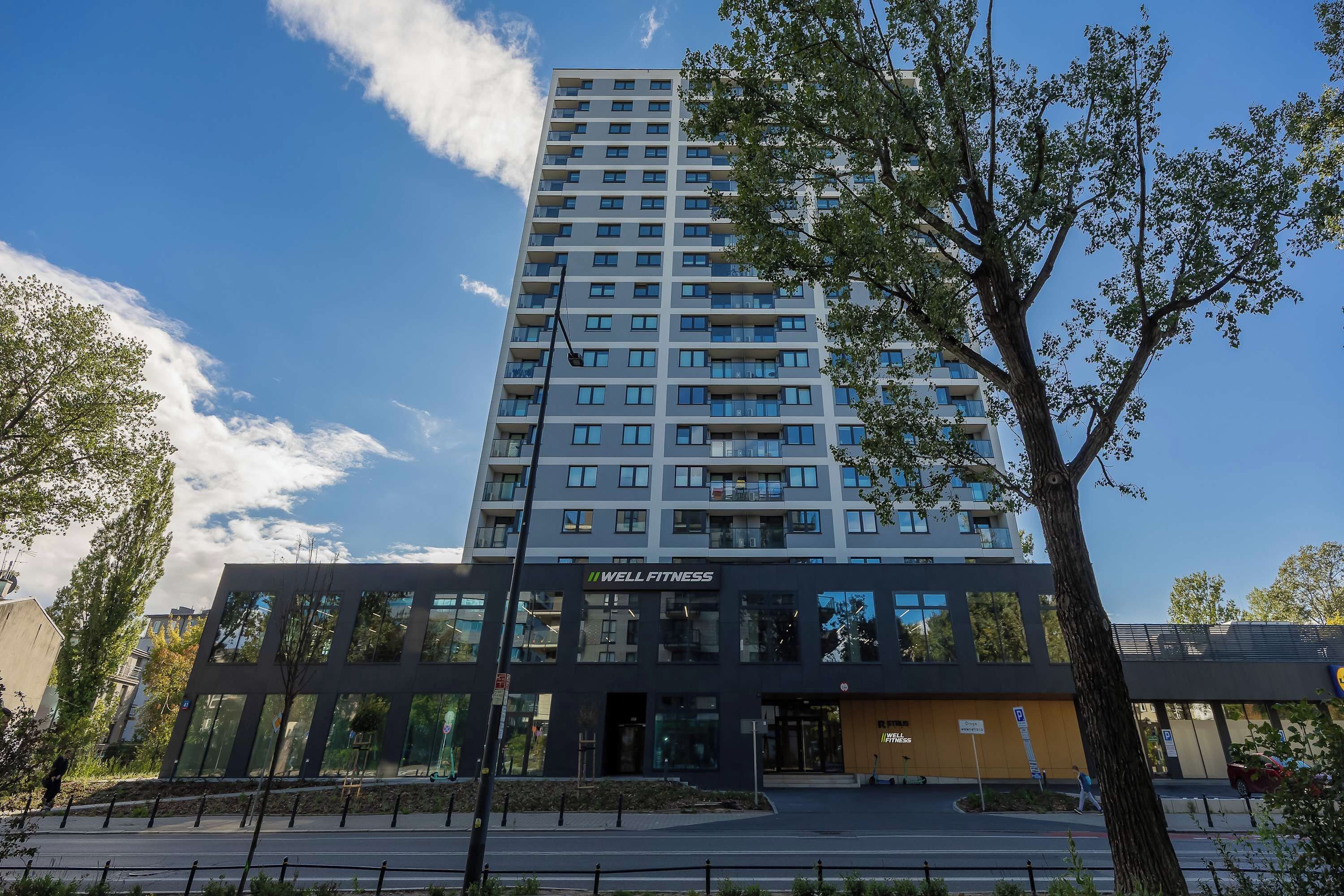 A modern multi-story building with a prominent 'WELL FITNESS' sign, surrounded by trees and a clear sky.