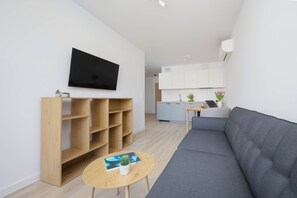 A modern living room with a flat-screen TV, a grey sofa, a wooden shelving unit, and a small round table with a magazine and a plant. - Warsaw | Grenadierów 41 | Modern Apartment | Air Conditioning (Warszawa)