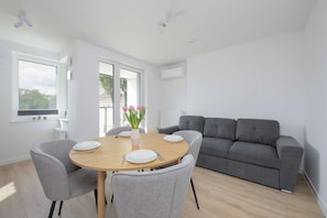A modern living room with a round wooden dining table, grey upholstered chairs, a grey sofa, and a window with a view of trees and buildings. - Warsaw | Grenadierów 41 | Comfortable Apartment | Air Conditioning (Warszawa)