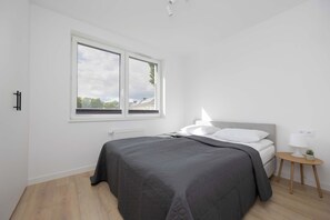 Un dormitorio moderno con un ventanal grande, una cama con ropa de cama gris, una mesita de noche con una lámpara y una pequeña planta.