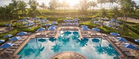 A pool area with lounge chairs and umbrellas, surrounded by palm trees and a view of the ocean.
