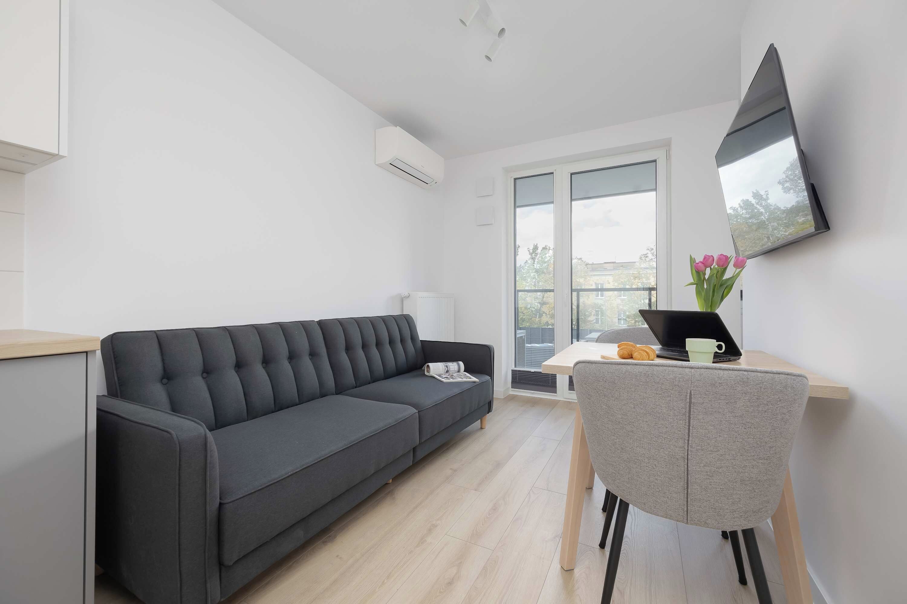 Una sala de estar moderna con un sofá gris oscuro, una mesita con una computadora portátil y una taza, y un ventanal con vistas al exterior.