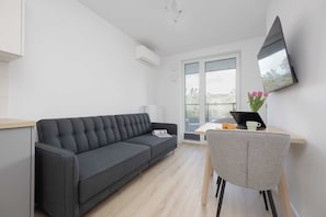 A modern living room with a dark grey sofa, a small table with a laptop and a cup, and a large window with a view of the outside. - Warszawa | Grenadierów 41 | Air Conditioned Studio | Desk (Warszawa)