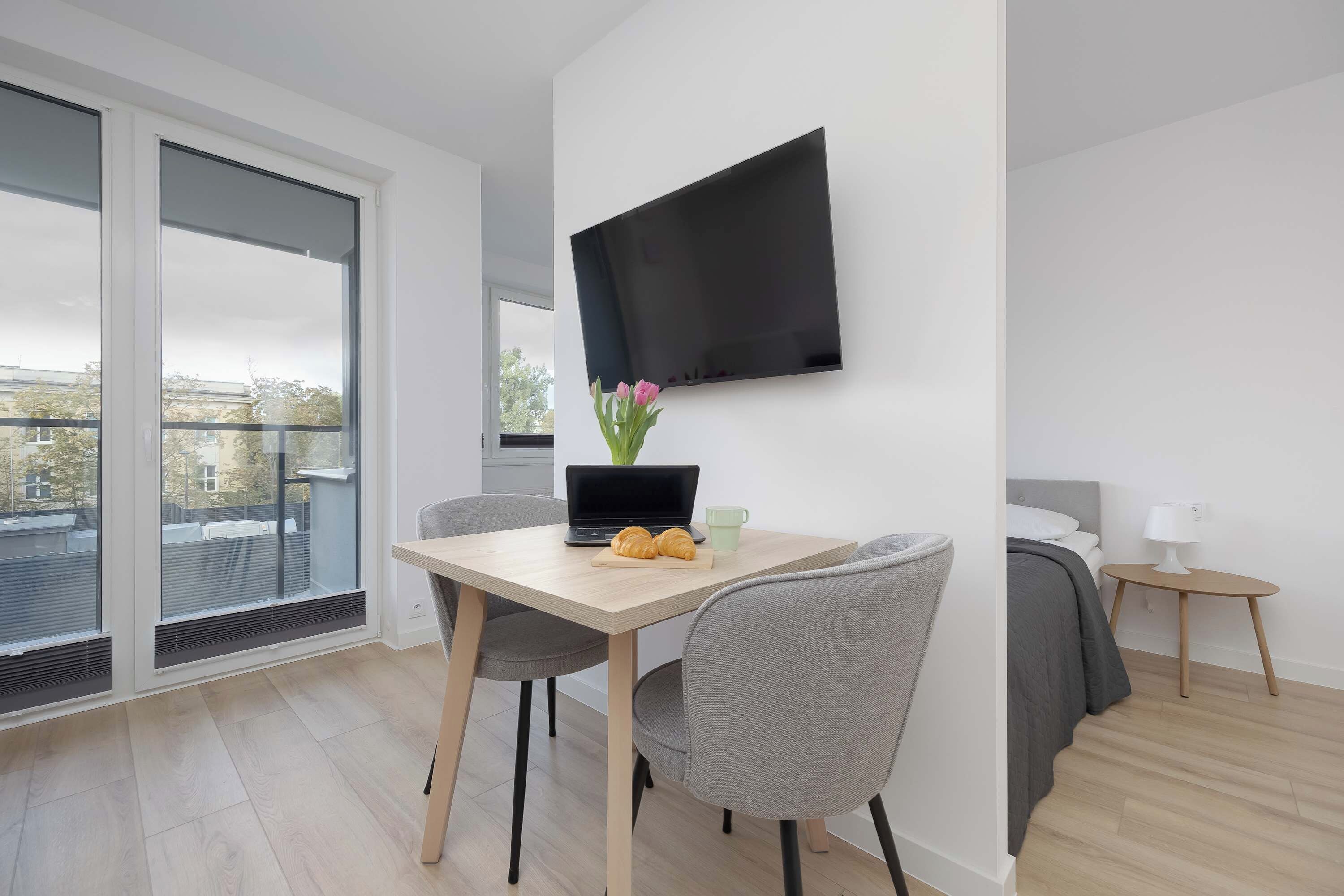A modern hotel room with a flat-screen TV mounted on the wall, a dining table with a laptop and croissants, a grey chair, and a bed with a grey headboard.