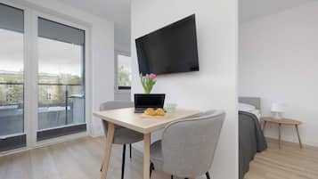 A modern hotel room with a flat-screen TV mounted on the wall, a dining table with a laptop and croissants, a grey chair, and a bed with a grey headboard.