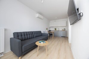 Una sala moderna con un sofá gris, una pequeña mesa de centro de madera y una zona de cocina con armarios blancos y un televisor de pantalla plana montado en la pared.