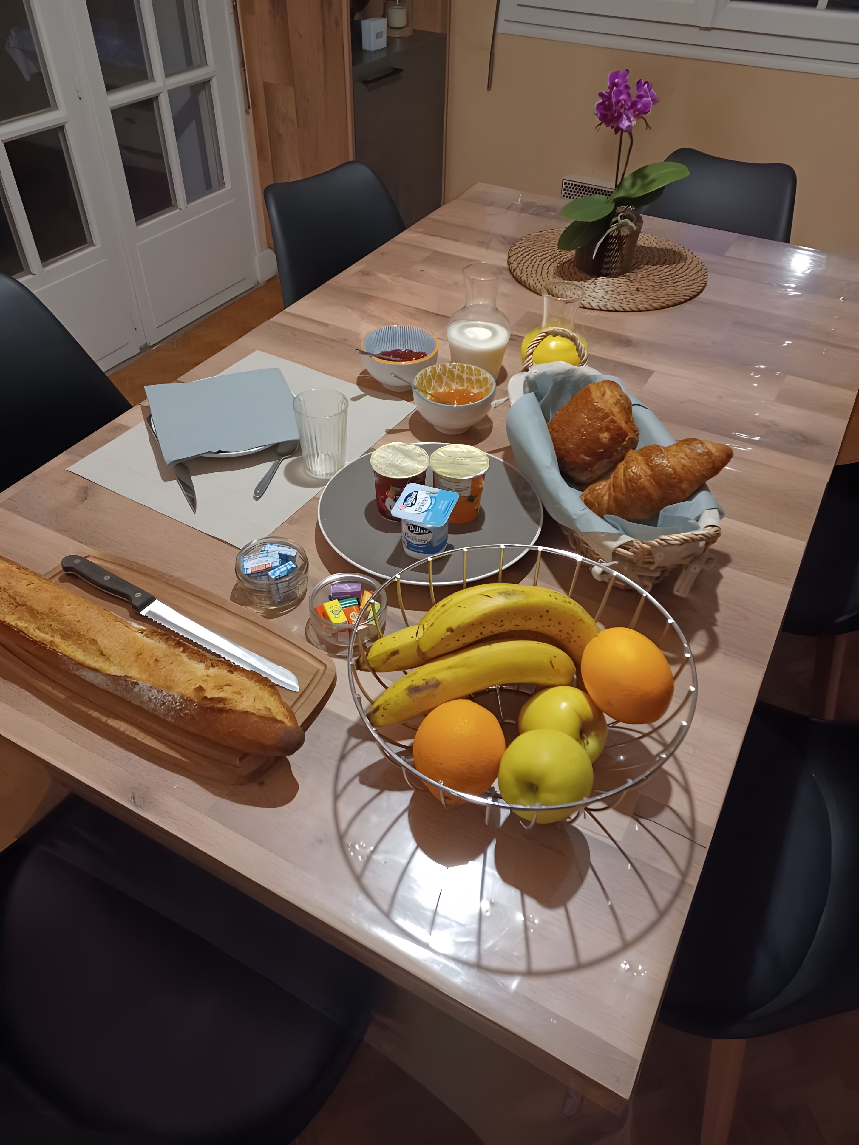 Une table à manger avec des plats, un petit pain, un panier de fruits et un vase rempli de fleurs.
