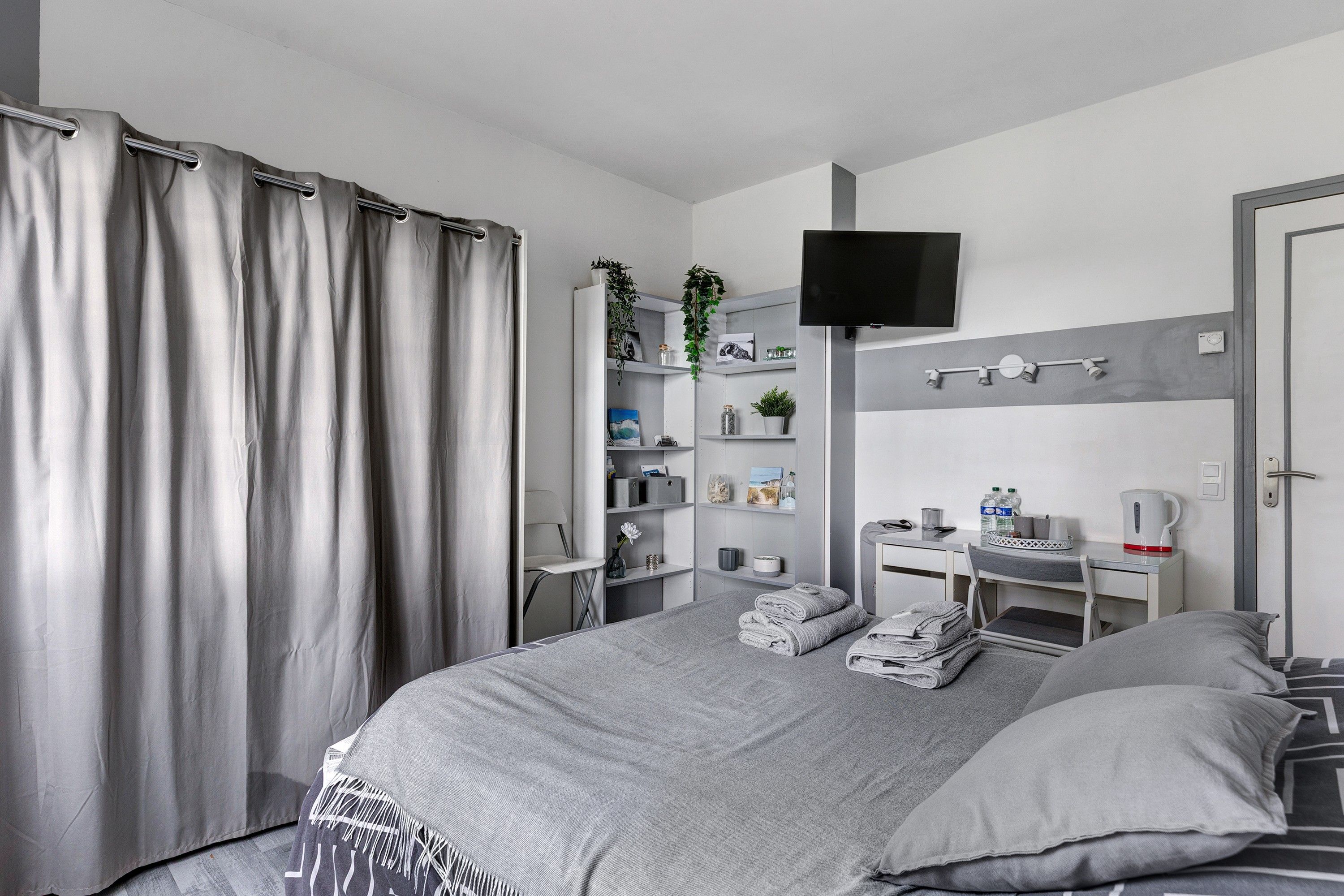 A modern bedroom with a bed, a TV, a desk, and a shelf with various items.