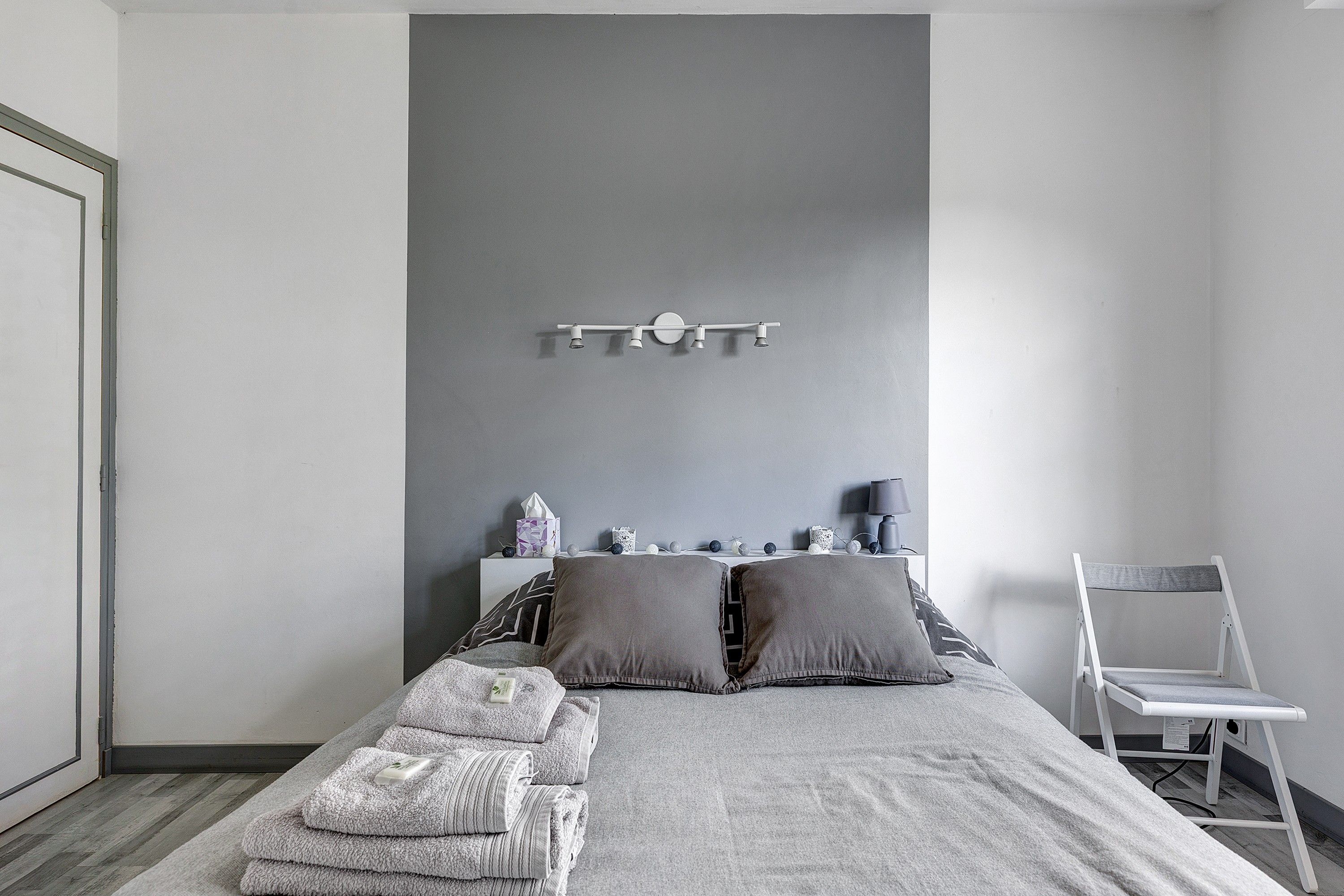 Een slaapkamer met een bed, kussens, een nachtkastje met een lamp en een stoel.