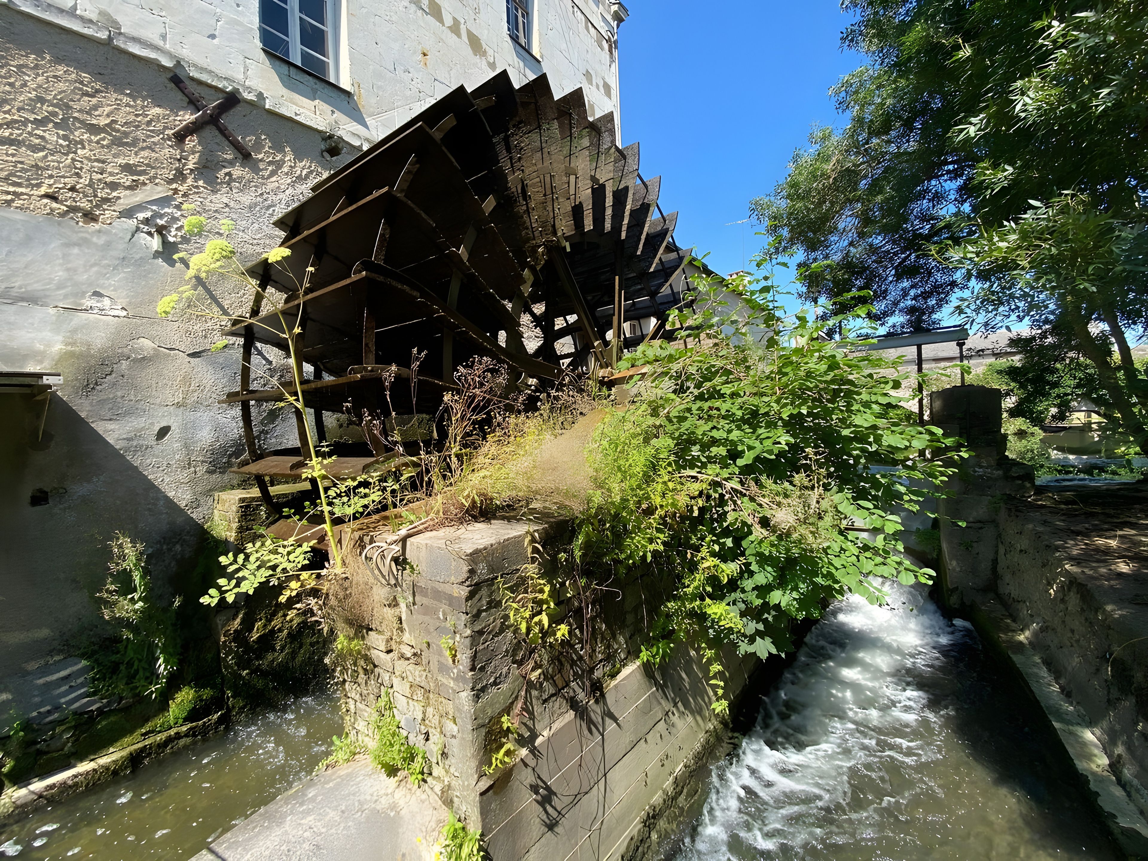 Une roue à eau historique, située à côté d’un bâtiment dont le mur est en pierre, et un petit ruisseau qui coule à proximité.