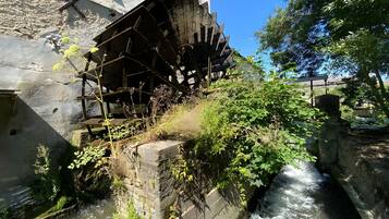 Une roue à eau historique, située à côté d’un bâtiment dont le mur est en pierre, et un petit ruisseau qui coule à proximité.
