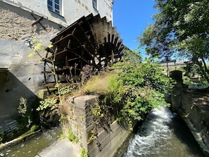 A historic water wheel structure adjacent to a building with a stone wall and a small stream flowing beside it.