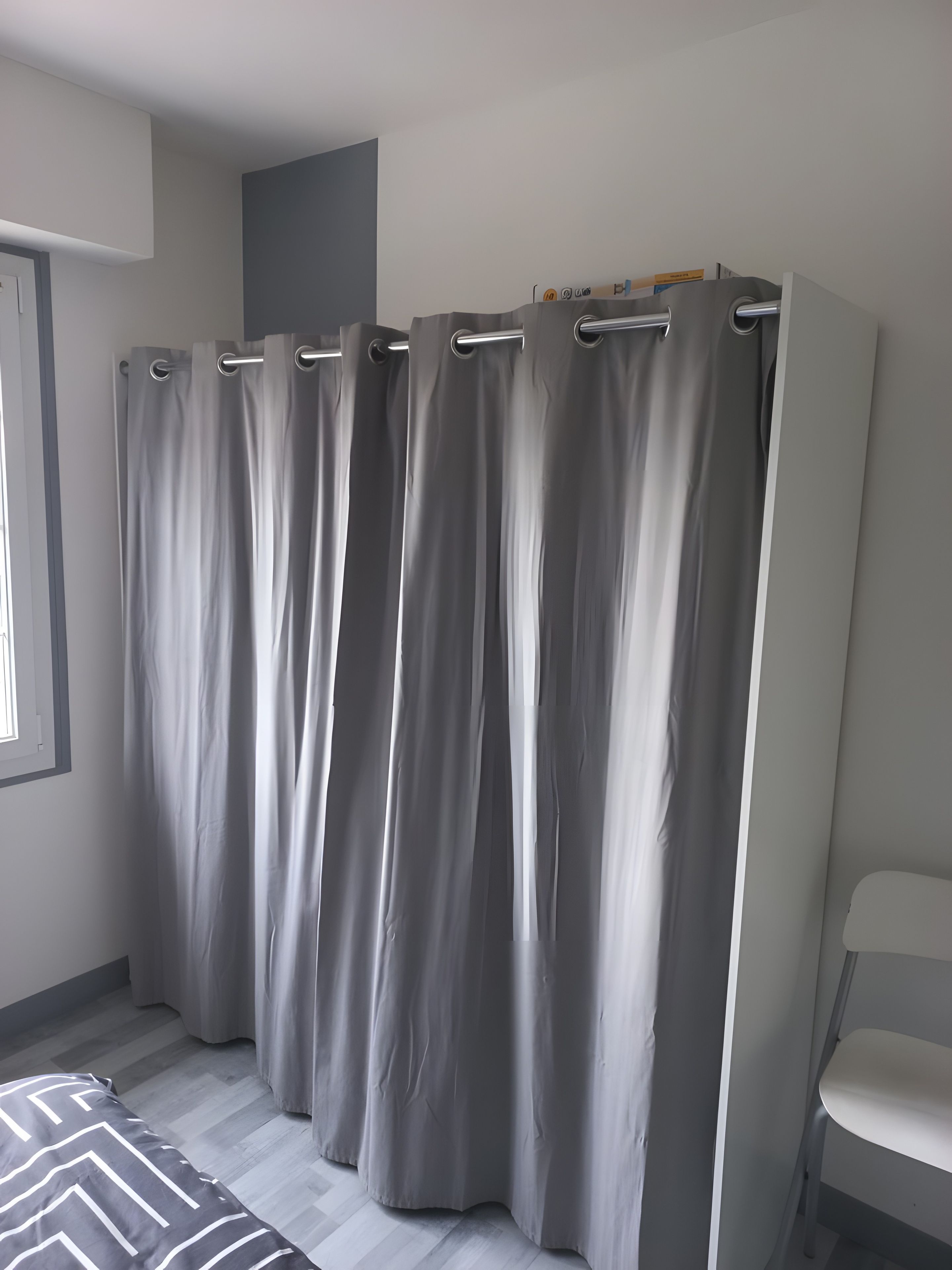 Une pièce avec un rideau gris, un lit recouvert d’une couvre-lit à motifs et une chaise blanche.