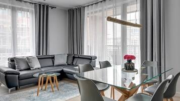 A modern living room with a glass dining table, grey sofa, and a large window with sheer curtains.