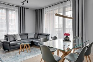 A modern living room with a glass dining table, grey sofa, and a large window with sheer curtains. - Premium Apartment Mustang with Parking (Gdańsk)