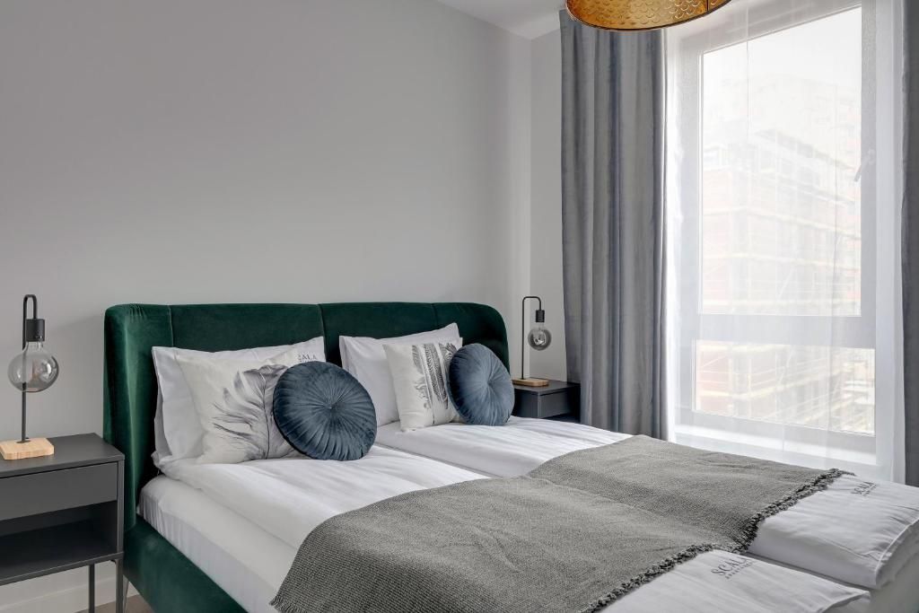 A modern bedroom with a green upholstered headboard, a white bedspread, and a grey throw blanket. There is a bedside table with a lamp and a small wooden block.