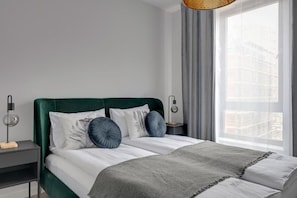 Een moderne slaapkamer met een groen gestoffeerd hoofdeinde, een witte spreideken en een grijze plaid. Er staat een nachtkastje met een lamp en een klein houten blokje.