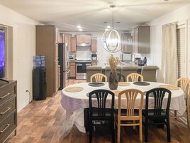 Comedor con una mesa lista para cuatro personas, rodeada de sillas, en una habitación con piso de madera y una cocina al fondo.