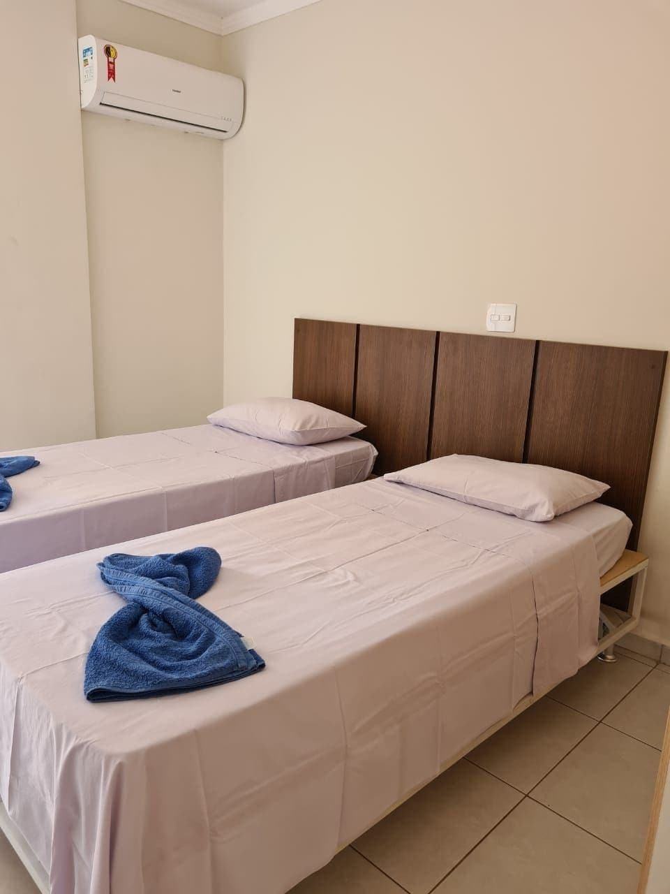 Quarto de hotel com duas camas de solteiro, cabeceira de madeira, ar condicionado e pequena mesa de cabeceira.