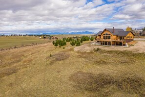 A large wooden house with a porch, surrounded by a grassy field and trees, with mountains in the background. - Big Bear Retreat (Gallatin Gateway)