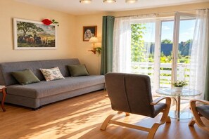 Ein Wohnzimmer mit einer Couch, einem Sessel und einem Tisch mit einer Pflanze. Ein Fenster mit Vorhängen bietet Blick auf Grünfläche.