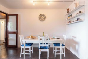 Un coin repas avec une table blanche, des chaises bleues à coussins, une assiette décorative accrochée au mur et une étagère blanche où sont disposés divers objets.