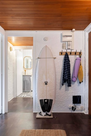A room with a surfboard leaning against the wall, a coat rack with items, and a door leading to another room. - Appt Bourdaines - Seignosse - Cozy - Beach 2min (Seignosse)
