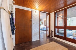 Une porte en bois avec un mur à texture, un couloir menant à une pièce où se trouvent une chaise blanche et une table, et une vue sur un espace extérieur à travers une porte vitrée.