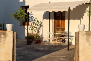 Eine Terrassengruppe im Freien mit Tisch und Stühlen, umgeben von Topfpflanzen und einem weißen Sonnensegel.