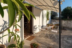 Eine Terrasse im Freien mit Tisch und Stühlen, umgeben von Blumentöpfen und einem überdachten Bereich.