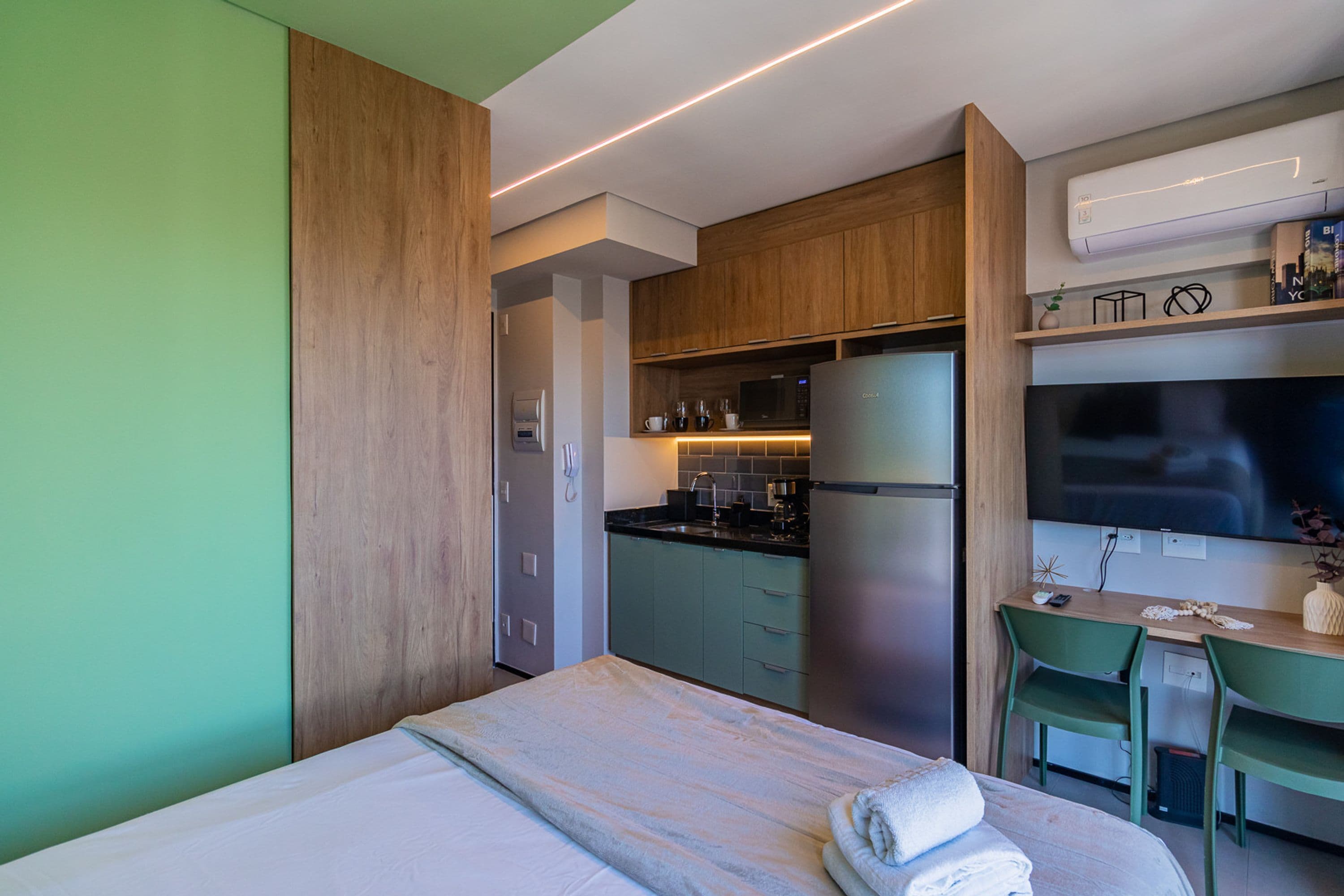 A compact hotel room with a bed, a kitchenette, a desk, and a TV.