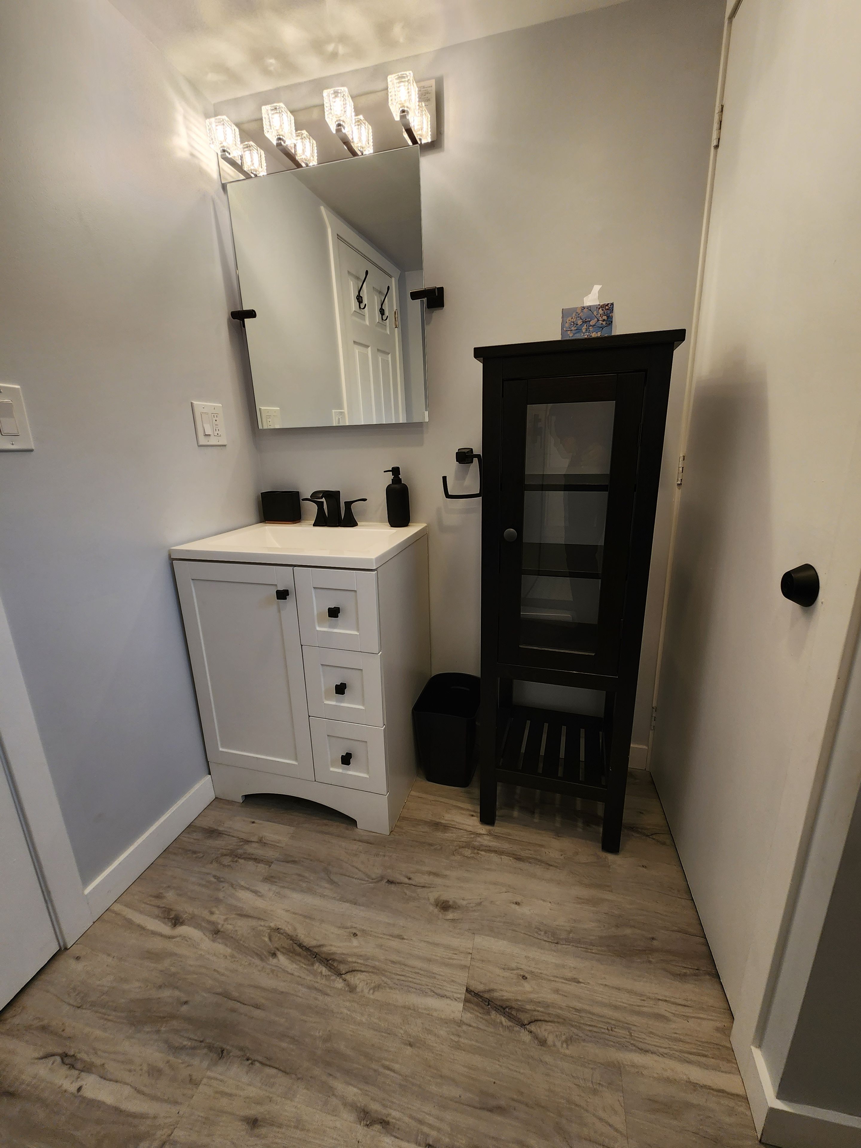A bathroom with a white vanity, a black cabinet, a mirror with lights, and a wooden floor.