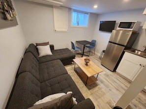 Un espace de vie compact comprenant un canapé, une petite table, un coin cuisine avec un réfrigérateur et un four à micro-ondes, et une fenêtre avec des stores.