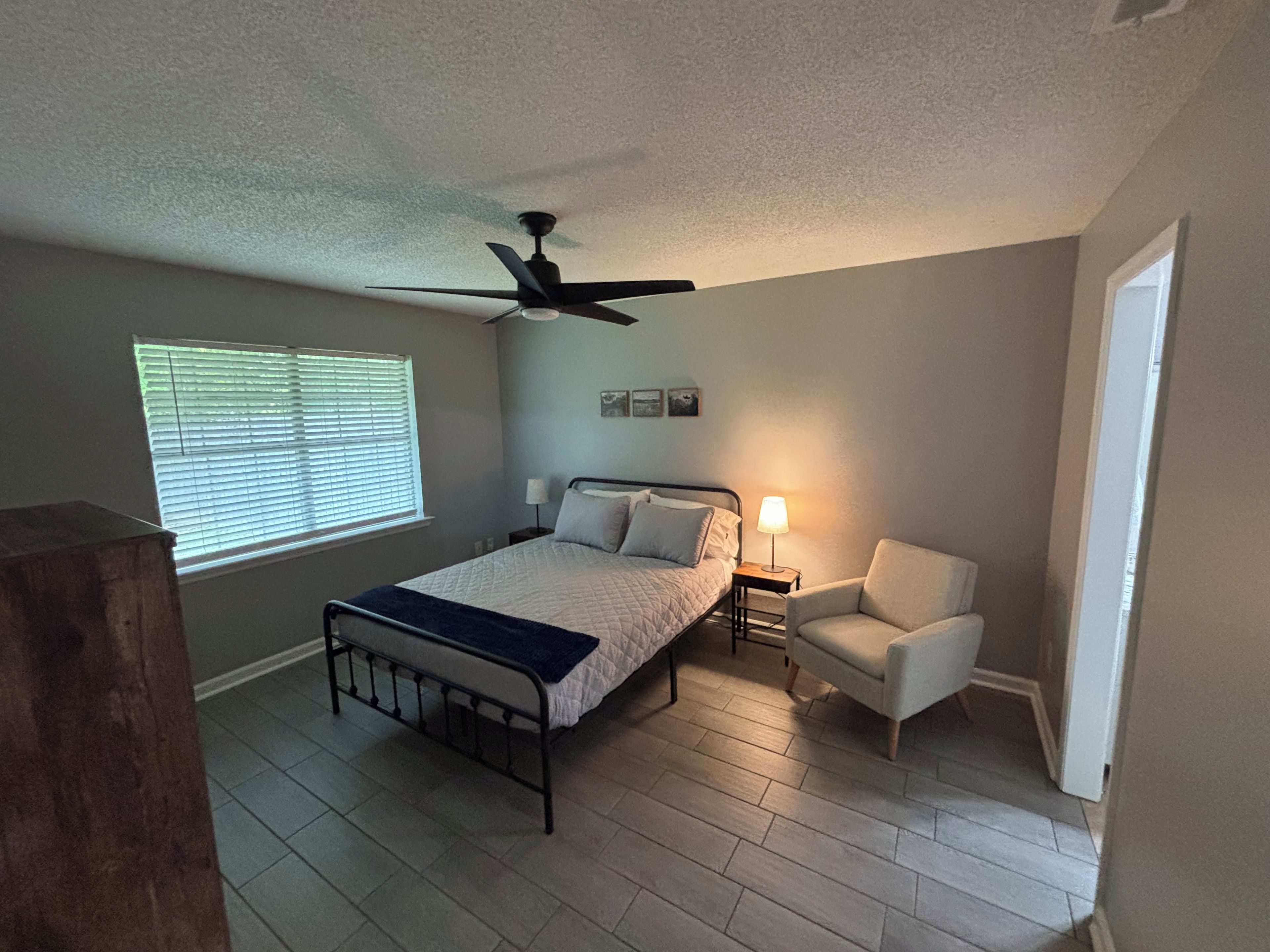 Un dormitorio con cama, una silla, una mesita de noche, ventilador de techo y una ventana con persianas.