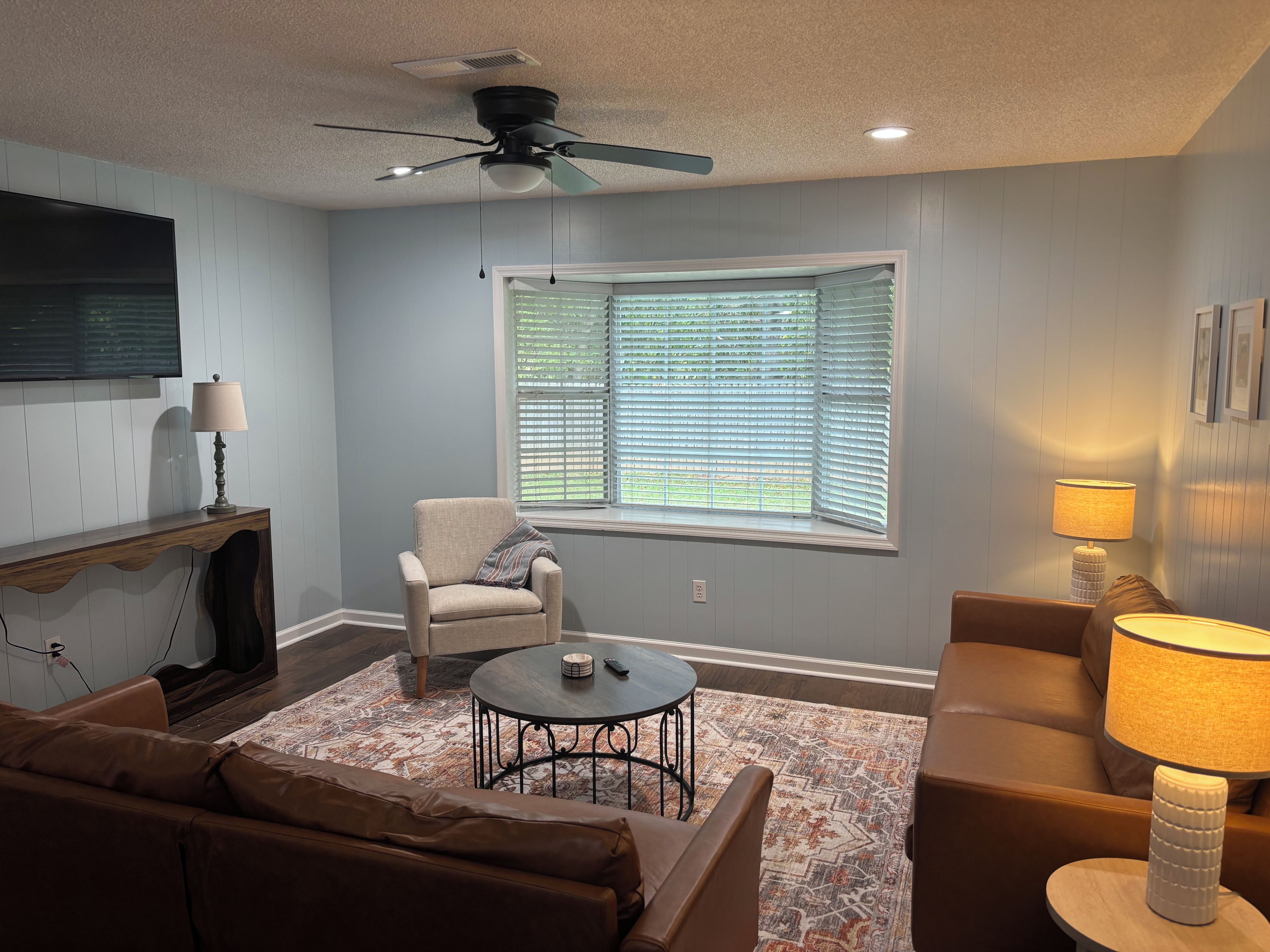 A living room with a ceiling fan, a window with blinds, a TV mounted on the wall, a sofa, a coffee table, and a lamp.