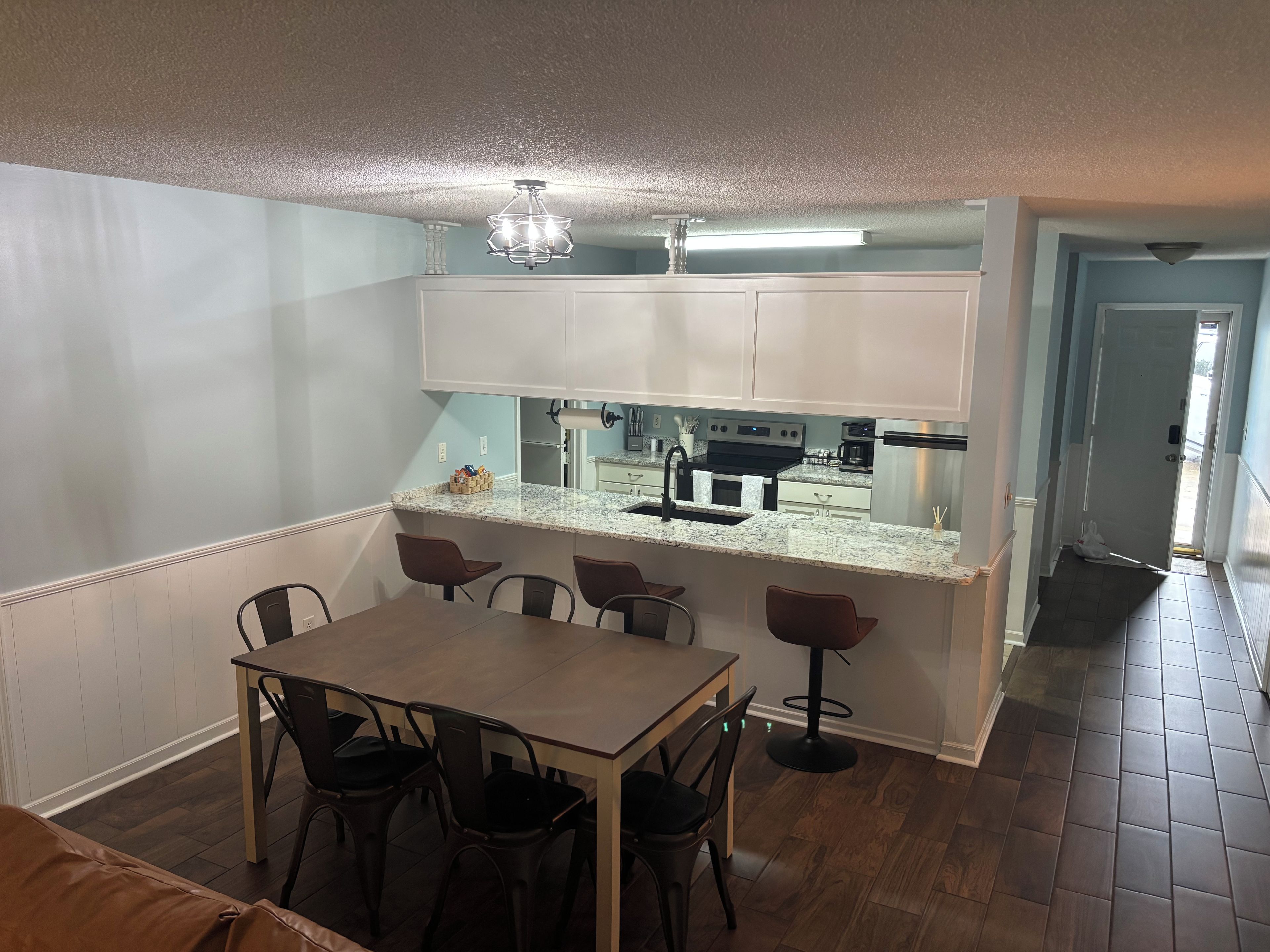 A dining area with a table and chairs, a kitchen with appliances, and a hallway leading to another room.