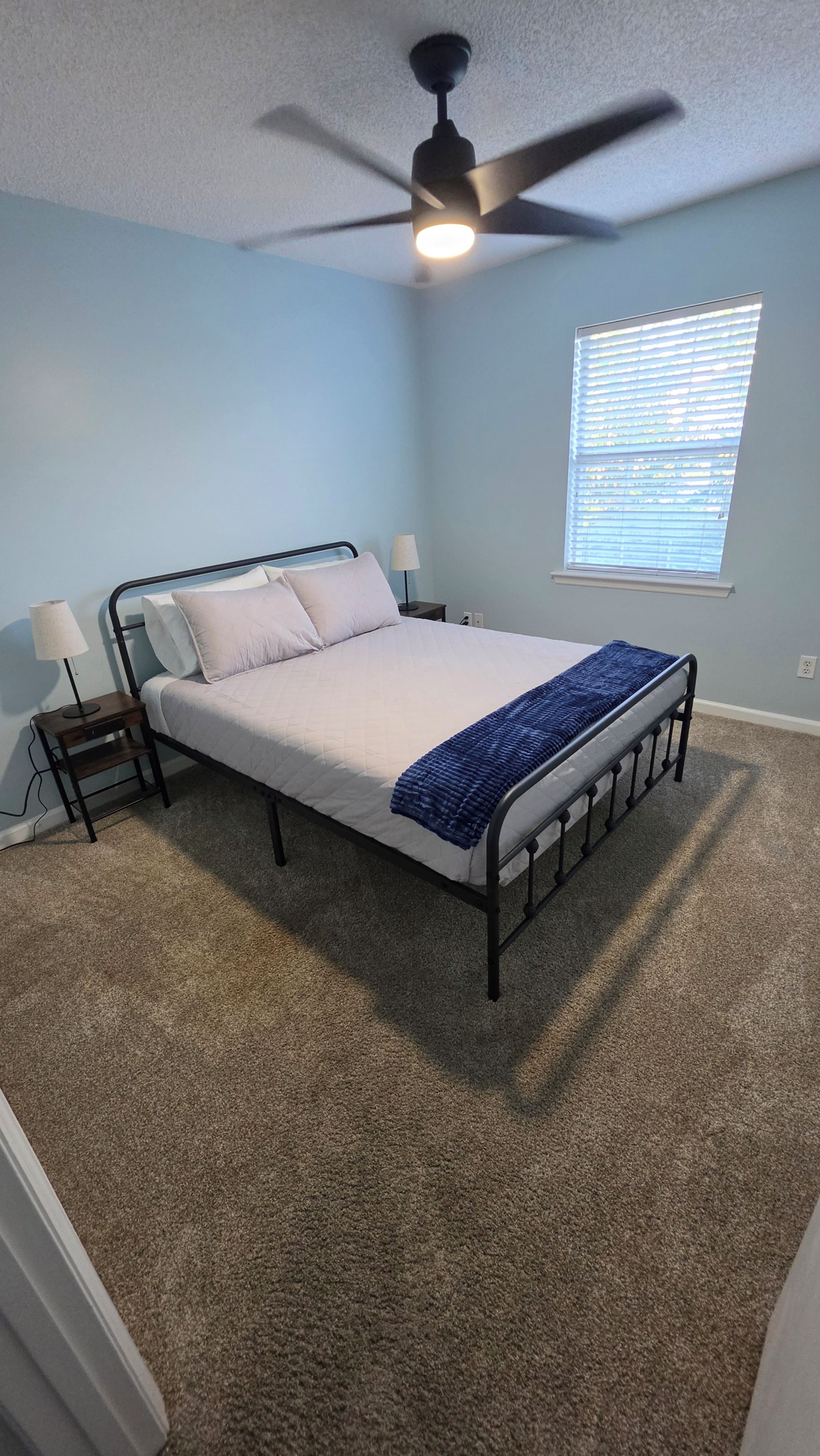A bedroom with a bed, a ceiling fan, a nightstand, a lamp, and a window with blinds.
