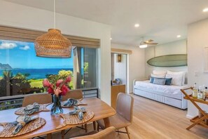 A modern room with a dining area, a bed with a view, and a large window overlooking the ocean. - Pali Ke Kua 124 - Bali Hai View, Beautiful Ocn Frt condo (Princeville)